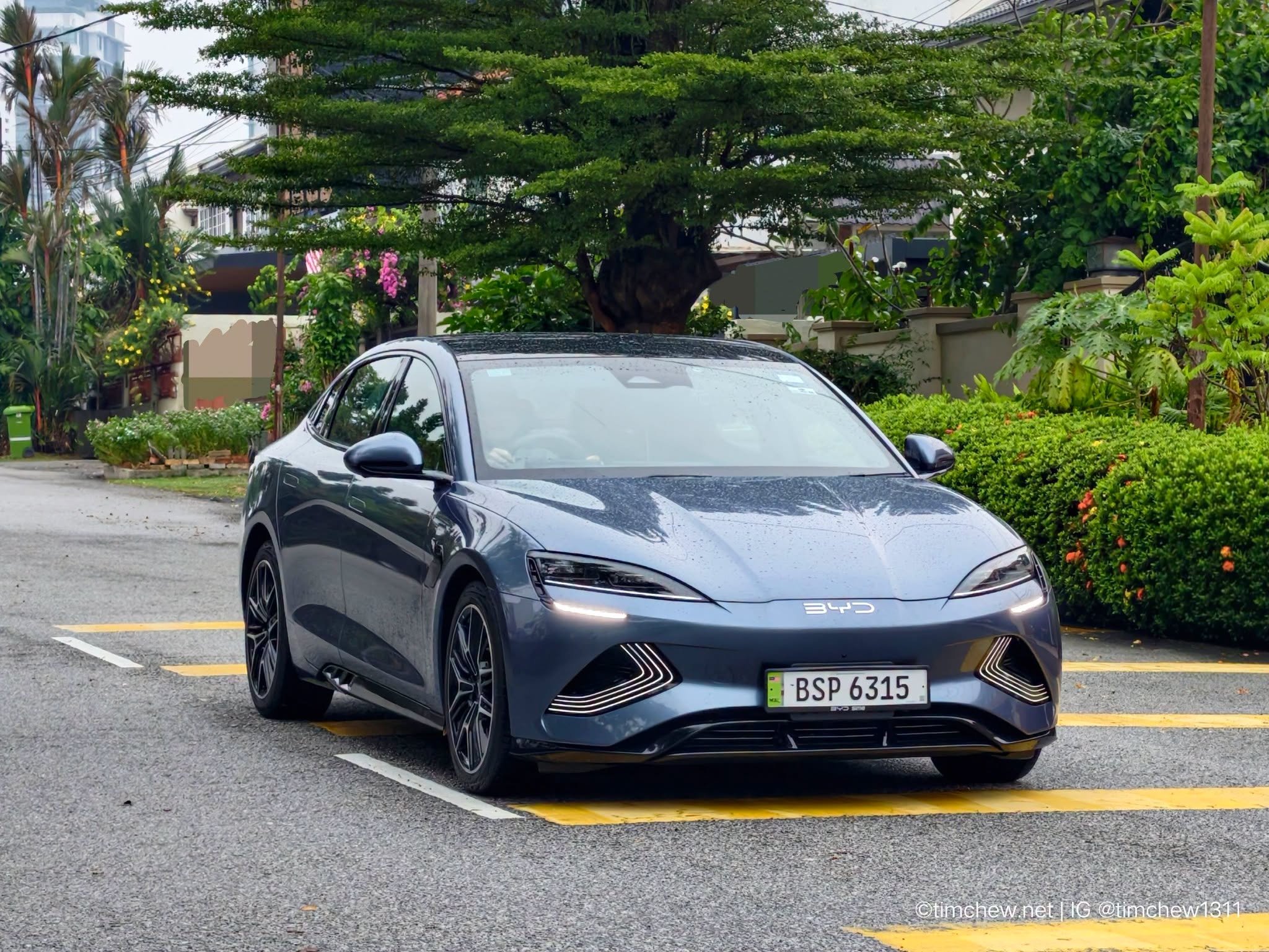 A sleek blue electric sedan parked on a street with greenery in the background.