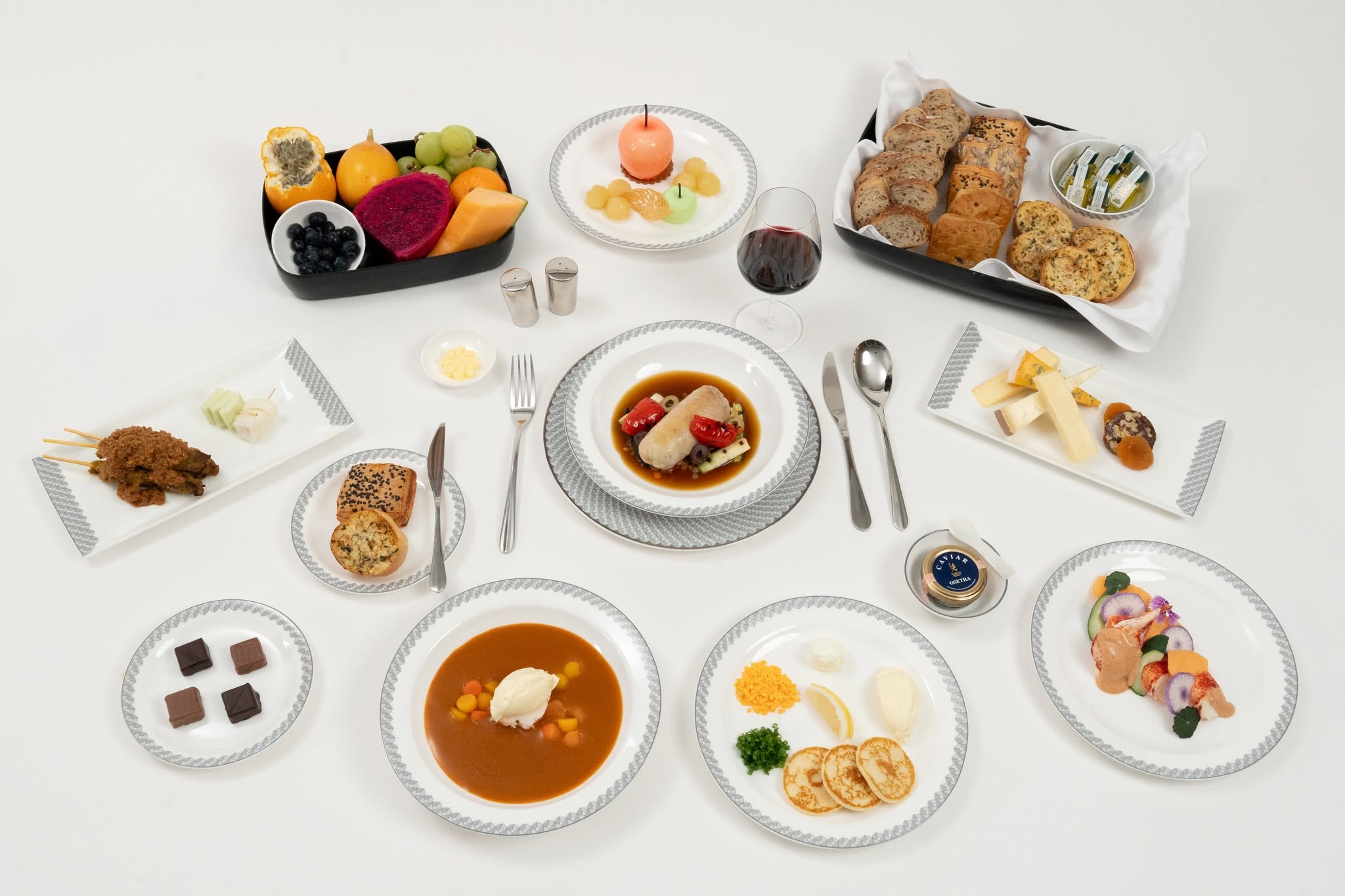 An assortment of gourmet dishes arranged on a white table, including satay skewers, fruit platter with various fruits, a bowl of soup with vegetables, dessert options like cakes and chocolates, and a centerpiece of main course on a decorative plate with sauce.