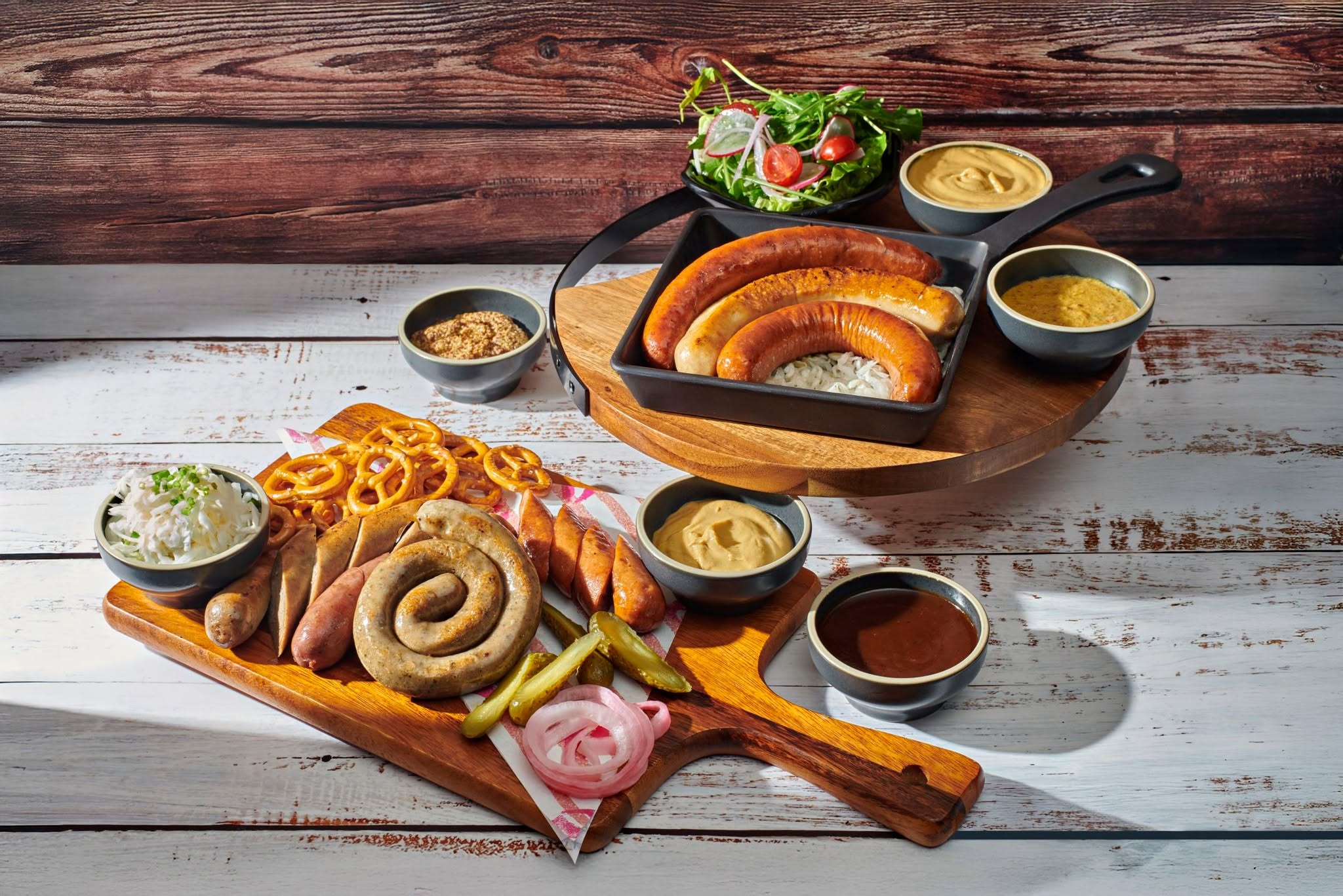 A wooden board featuring assorted sausages, pickles, and pretzels, accompanied by a bowl of chopped onions. In the background, a platter with grilled sausages, rice, a fresh salad, and several dipping sauces.