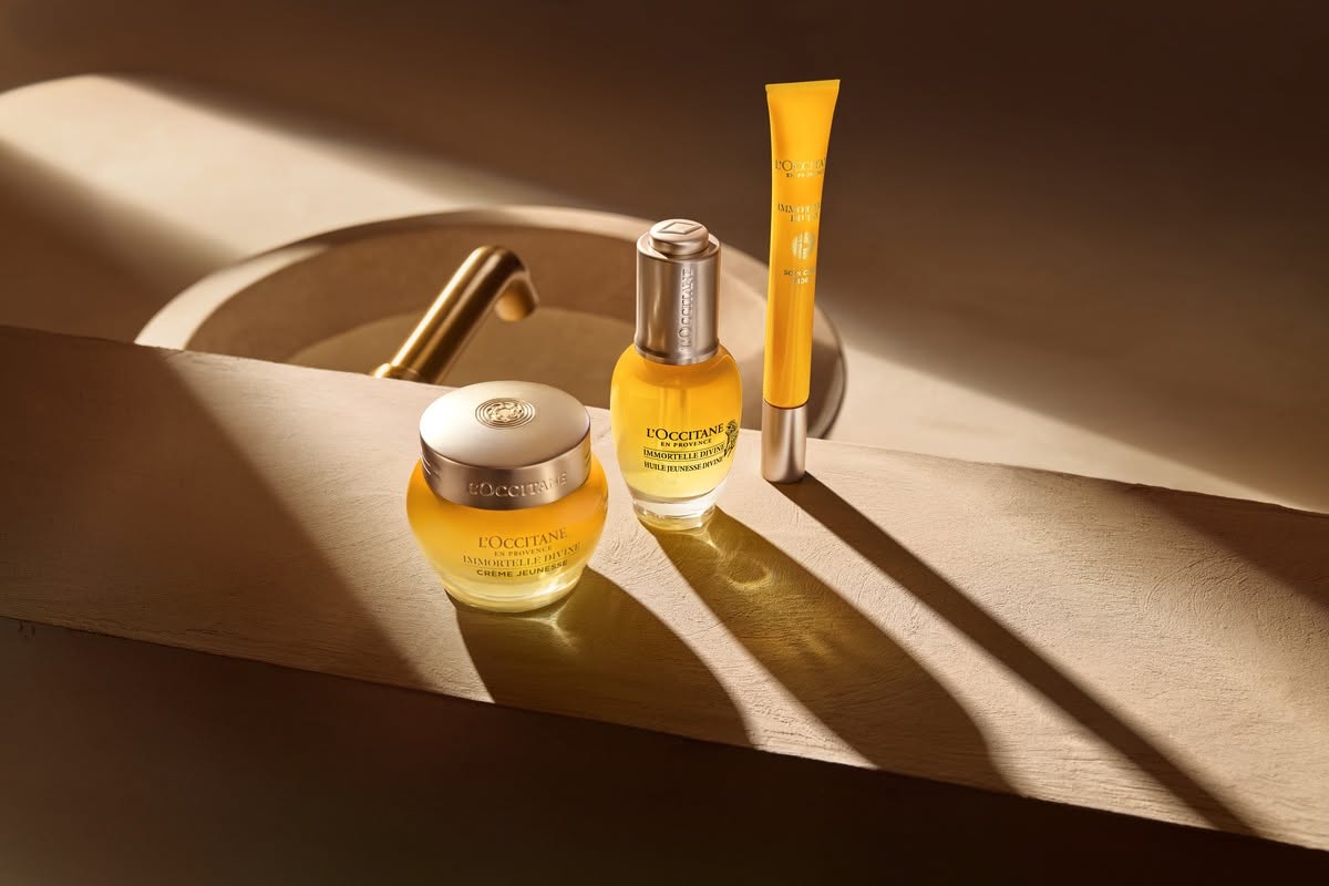 Three L'Occitane skincare products displayed on a wooden surface, featuring two yellow bottles and a tube, with soft sunlight casting shadows.