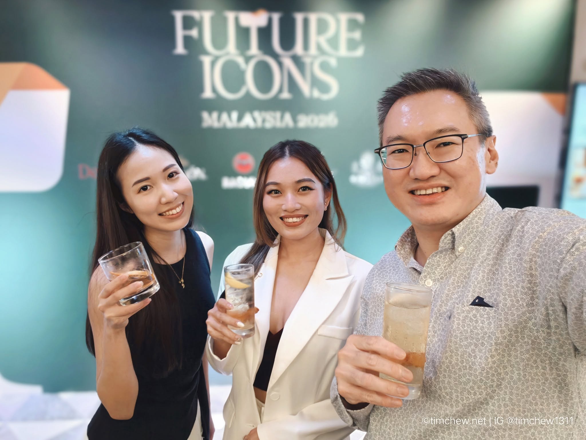 Three people smiling and posing for a selfie at an event, each holding a drink. The background features a banner that reads 'FUTURE ICONS MALAYSIA 2026'.