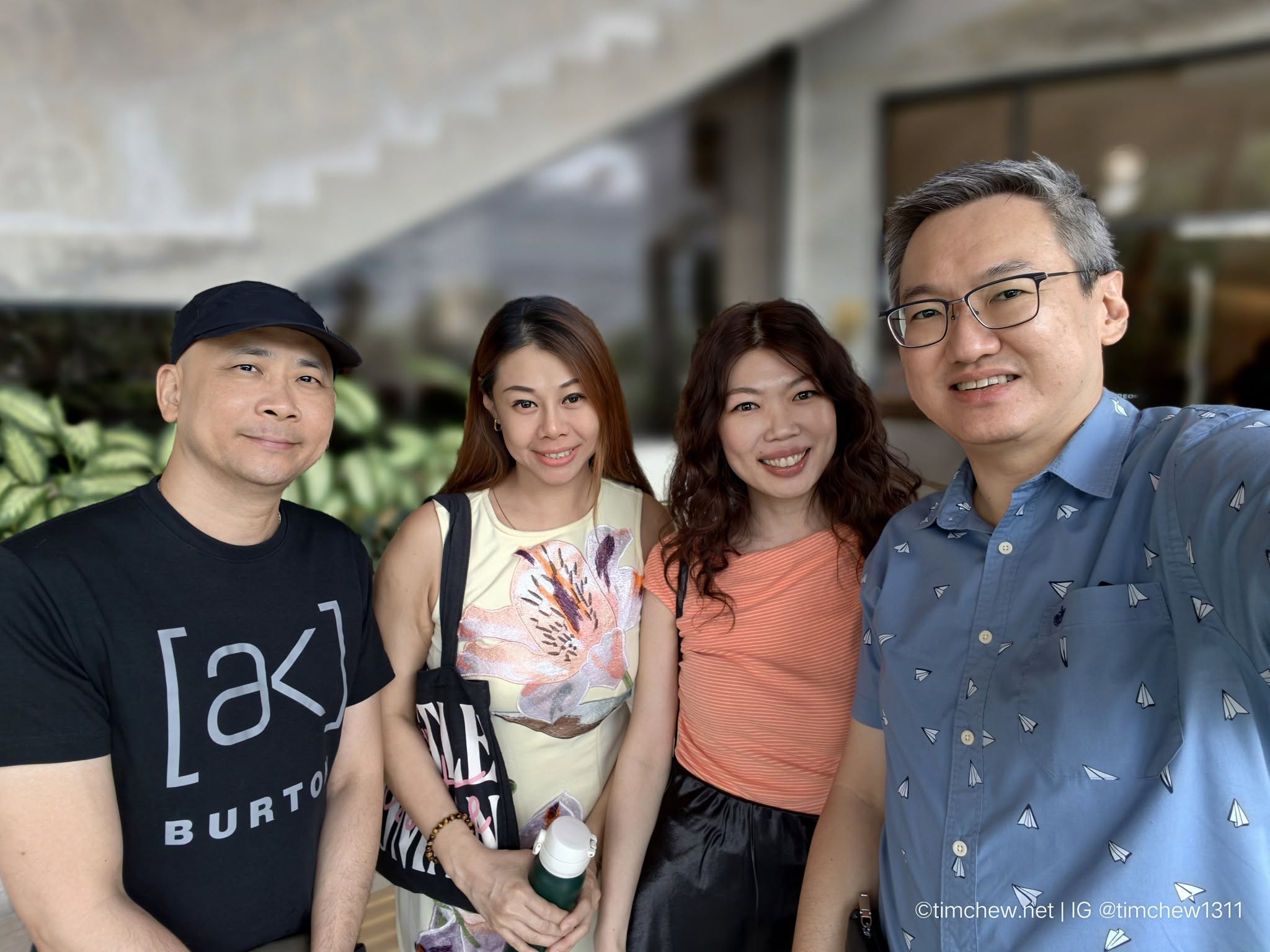 A group of four people posing for a photo outdoors. The first person on the left is wearing a black t-shirt and a cap. The second person is a woman in a floral top, next to her is another woman in an orange striped shirt, and the last person on the right is wearing a light blue patterned shirt. The background features greenery and architectural elements.