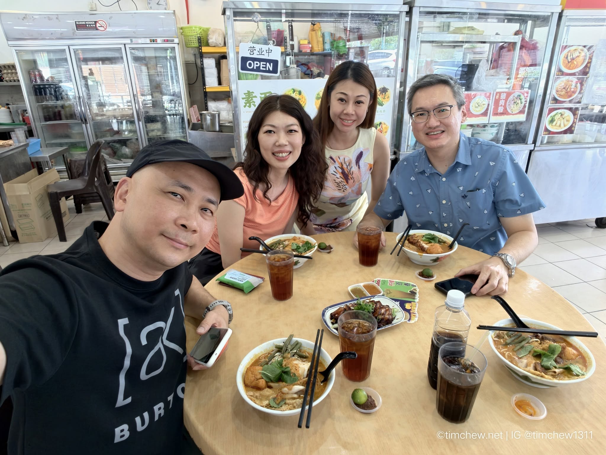 A group of four people enjoying a meal together at a restaurant table, featuring bowls of noodles and drinks in a casual eatery setting.