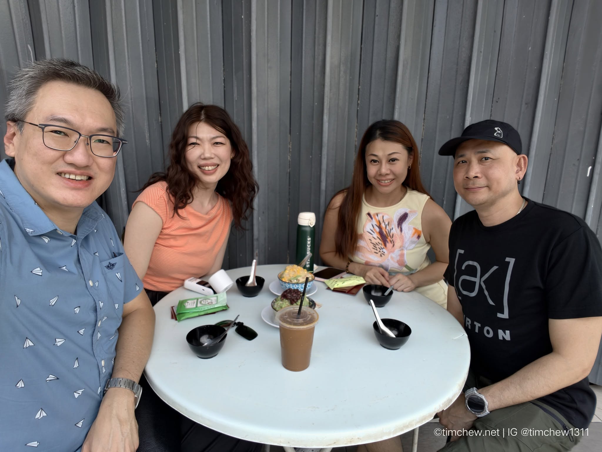 A group of four people sitting at a cafe table, enjoying snacks and drinks. The background features a textured gray wall.