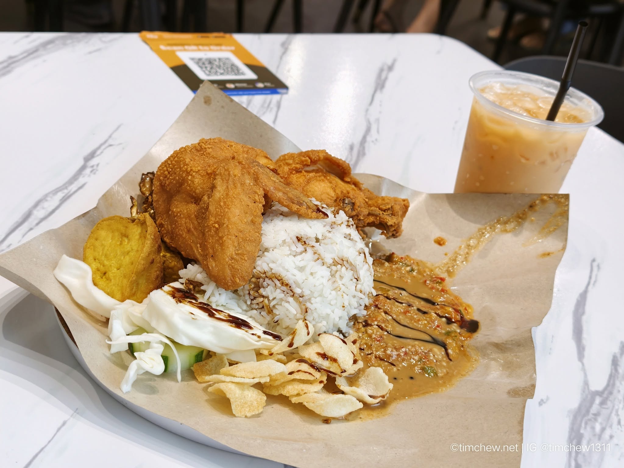 A plate of fragrant rice topped with crispy fried chicken, accompanied by golden fried potato, cucumber slices, a drizzle of sauce, and crispy snacks, served with an iced drink in the background.