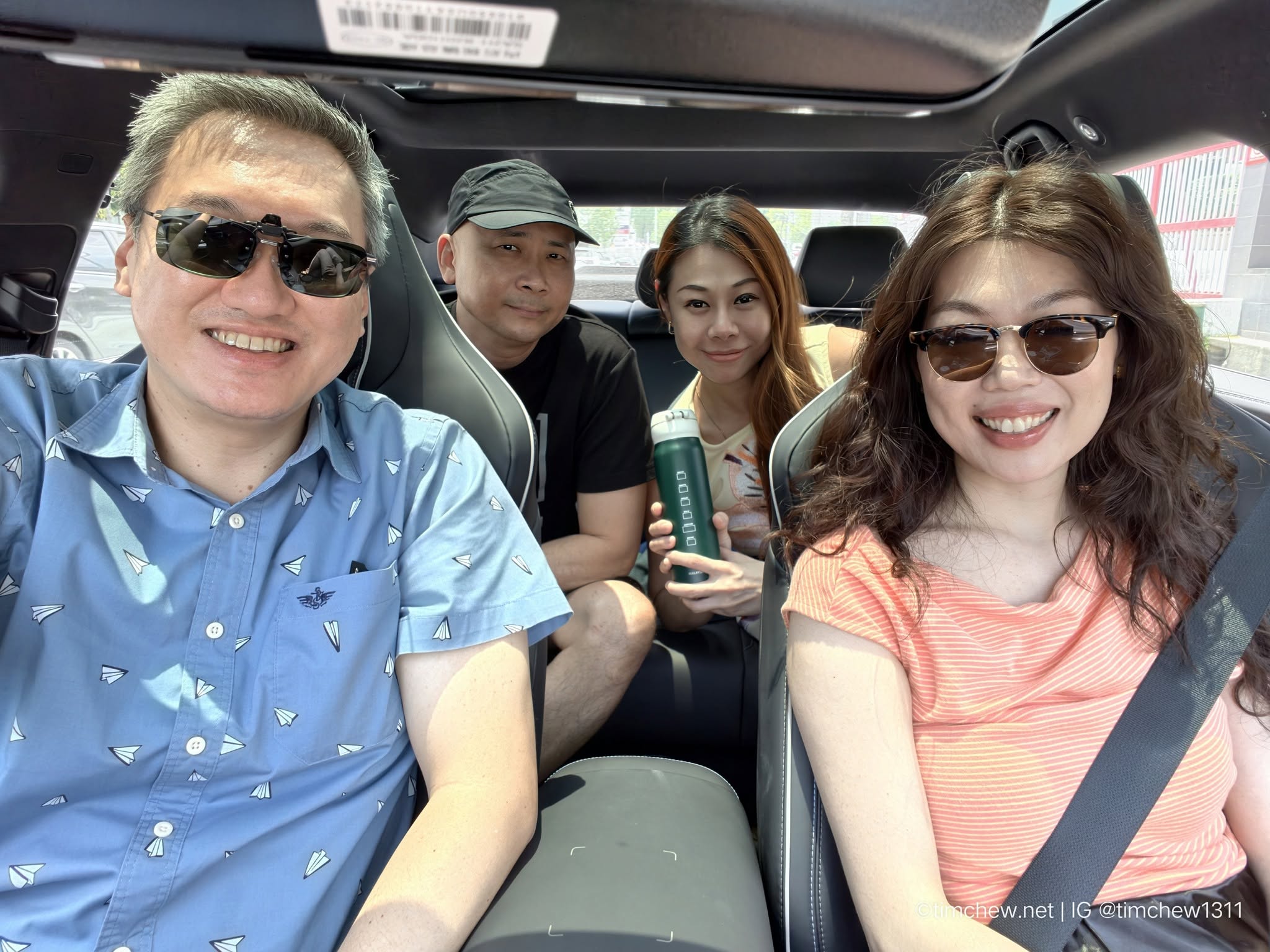 A group of four people smiling in a car, wearing sunglasses. Two men and two women are seated in the front and back seats, with one woman holding a green water bottle.