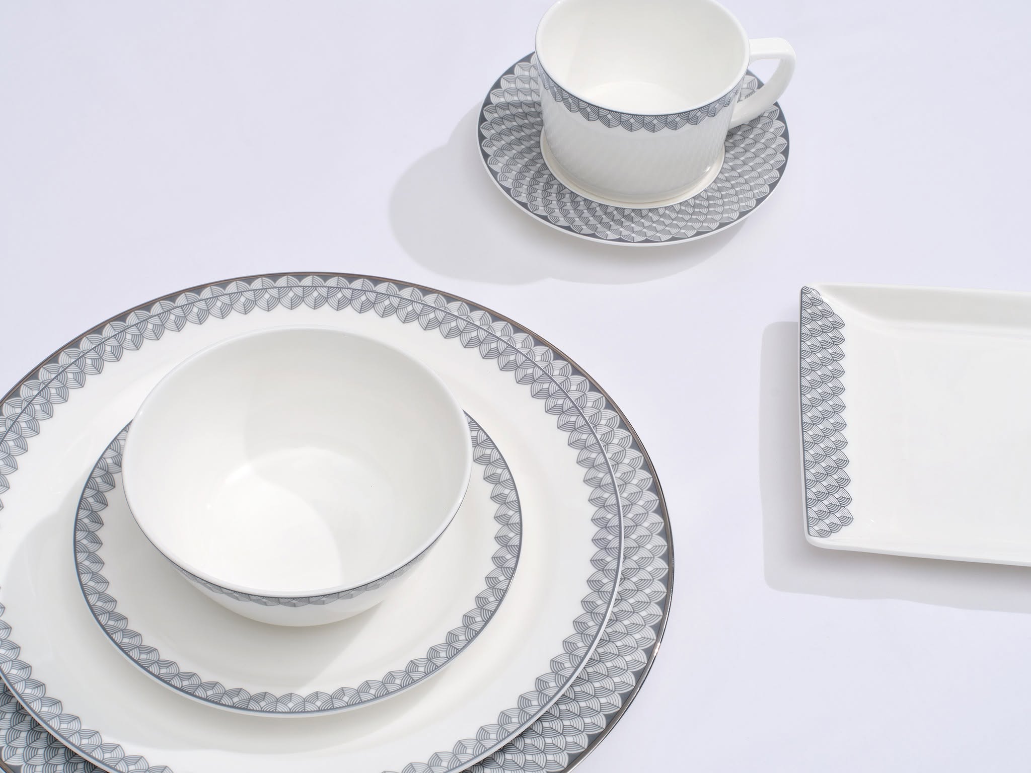 A collection of elegant white tableware featuring intricate gray geometric patterns, including a dinner plate, salad plate, bowl, cup, and saucer, arranged on a light background.
