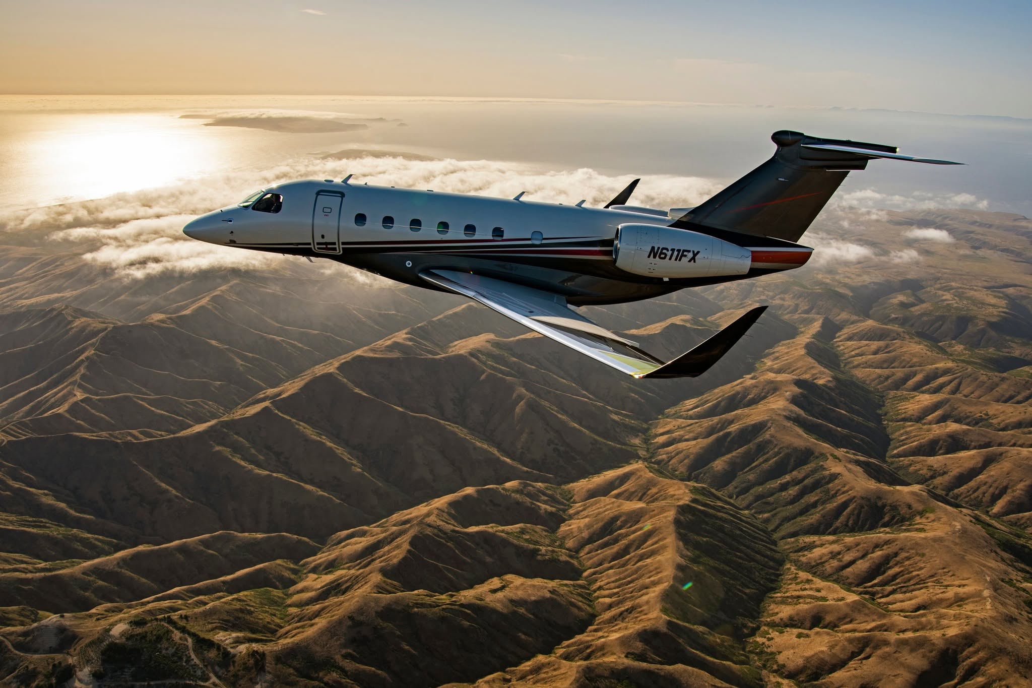 A sleek private jet flying over mountainous terrain with a sunset in the background.