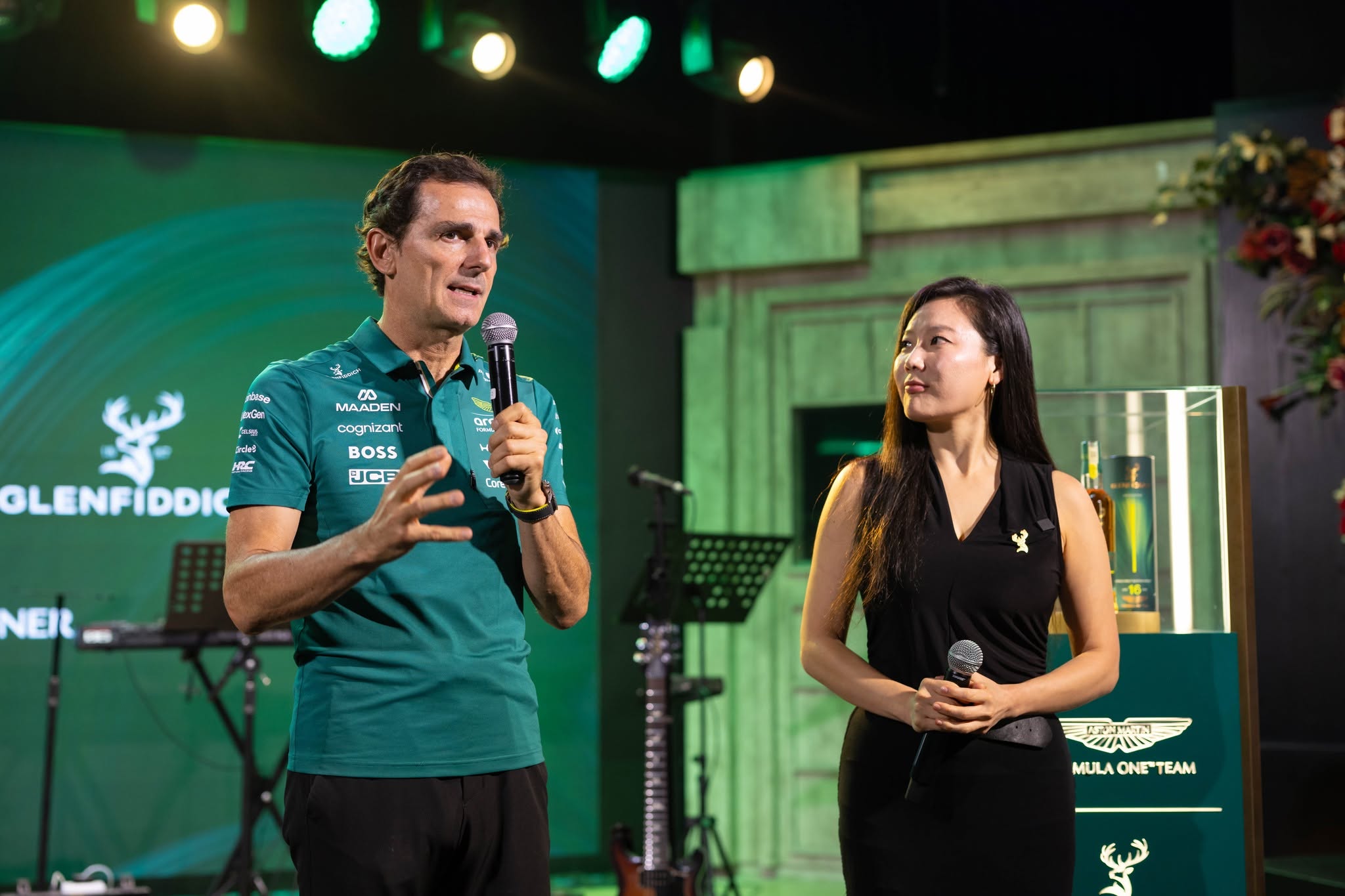A man in a green shirt speaking into a microphone while gesturing, with a woman in a black dress looking on. The background features a Glenfiddich logo and stage setup.