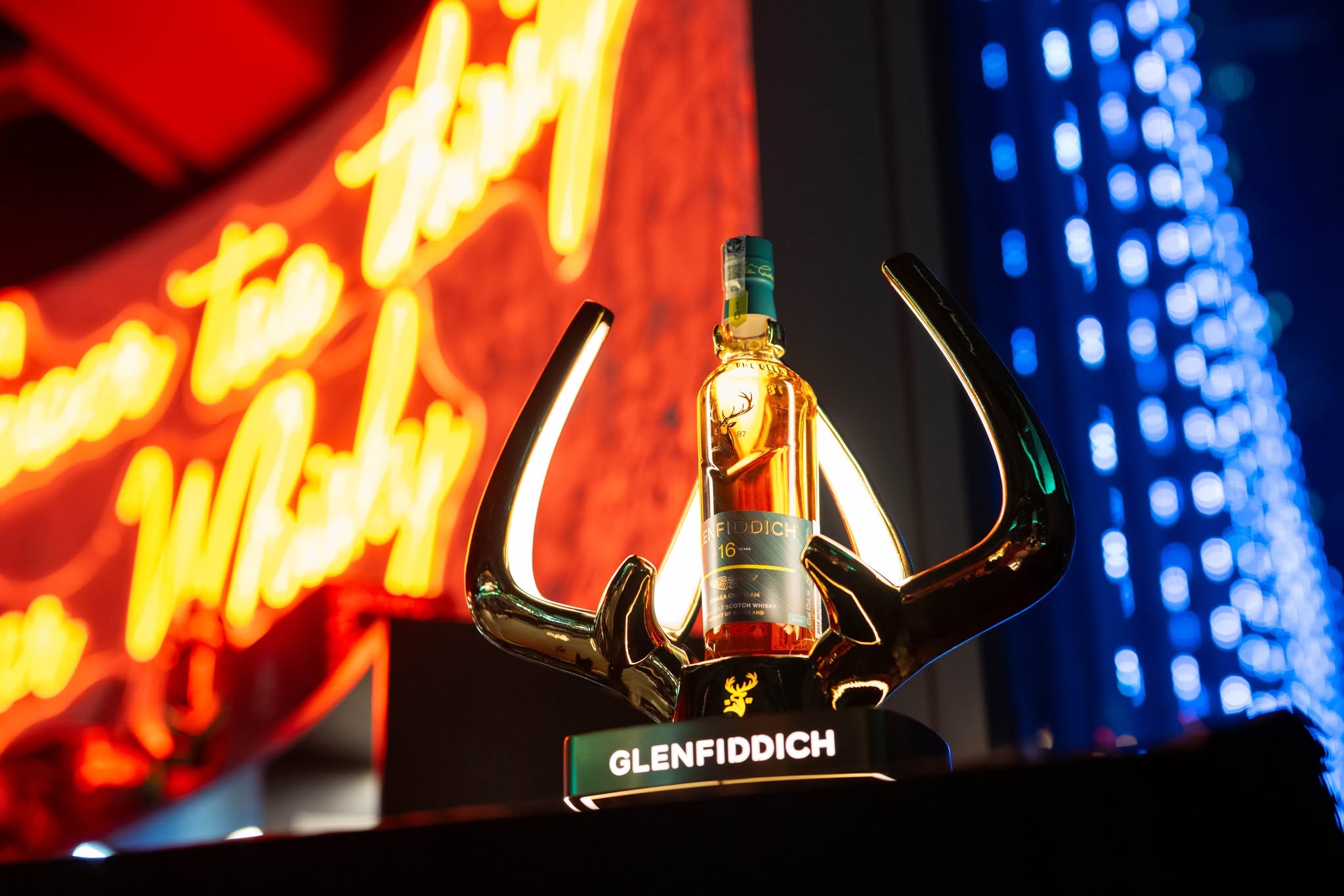 A bottle of Glenfiddich 16-year-old Scotch whisky displayed on a decorative antler stand, with a vibrant neon background.