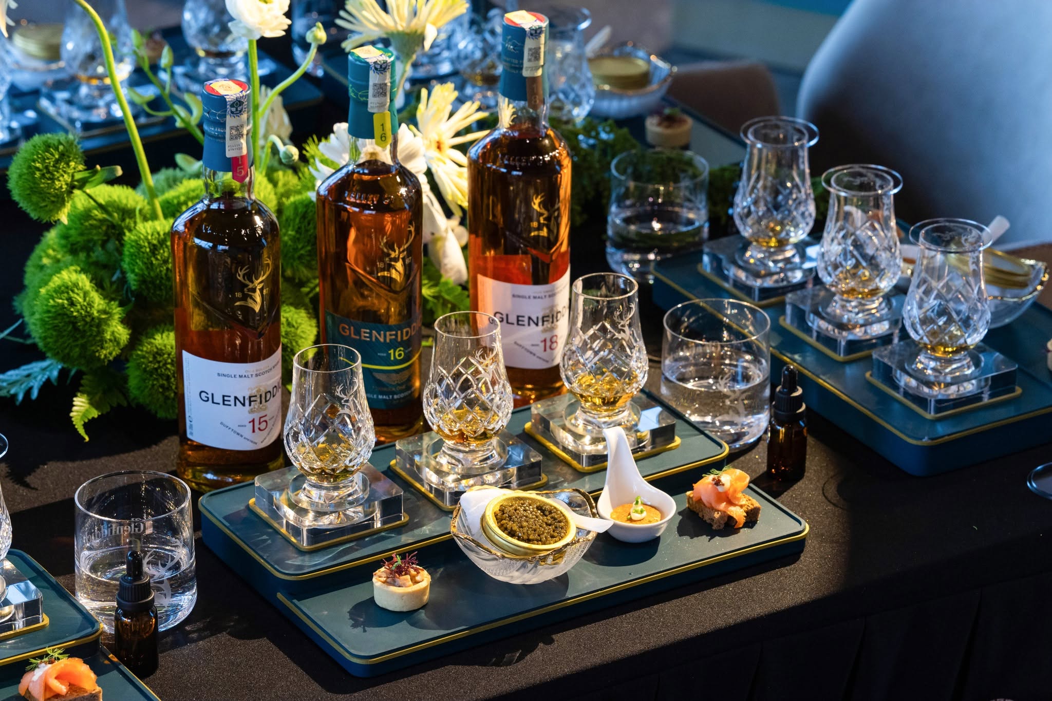 Table set with Glenfiddich whisky bottles, crystal glasses, and gourmet appetizers, surrounded by decorative greens.