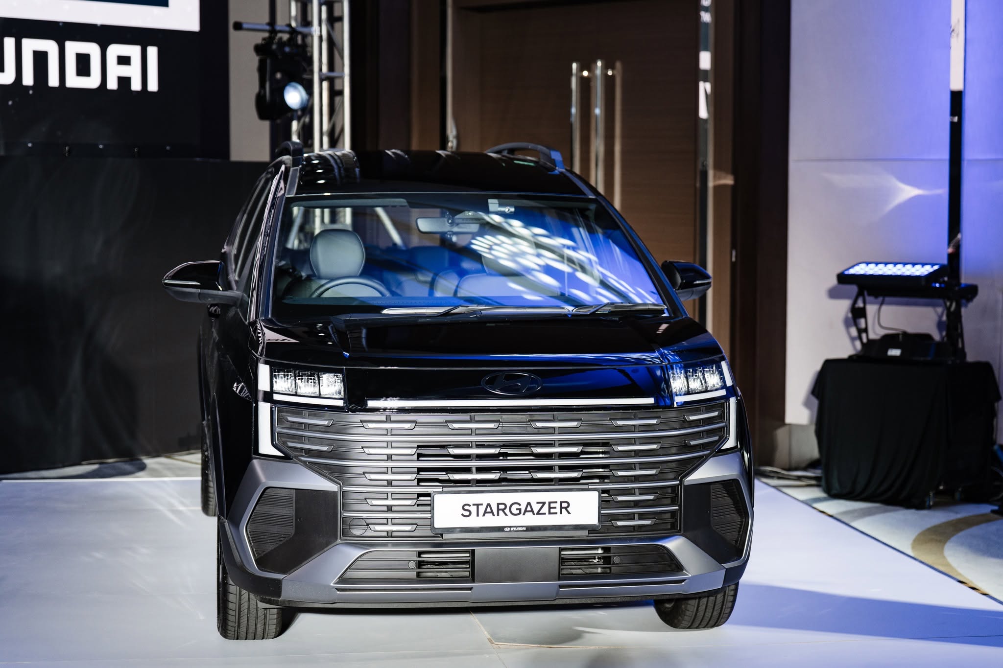 A front view of a black Hyundai Stargazer SUV, showcasing its sleek design and distinctive grille, set against a stylish indoor backdrop.
