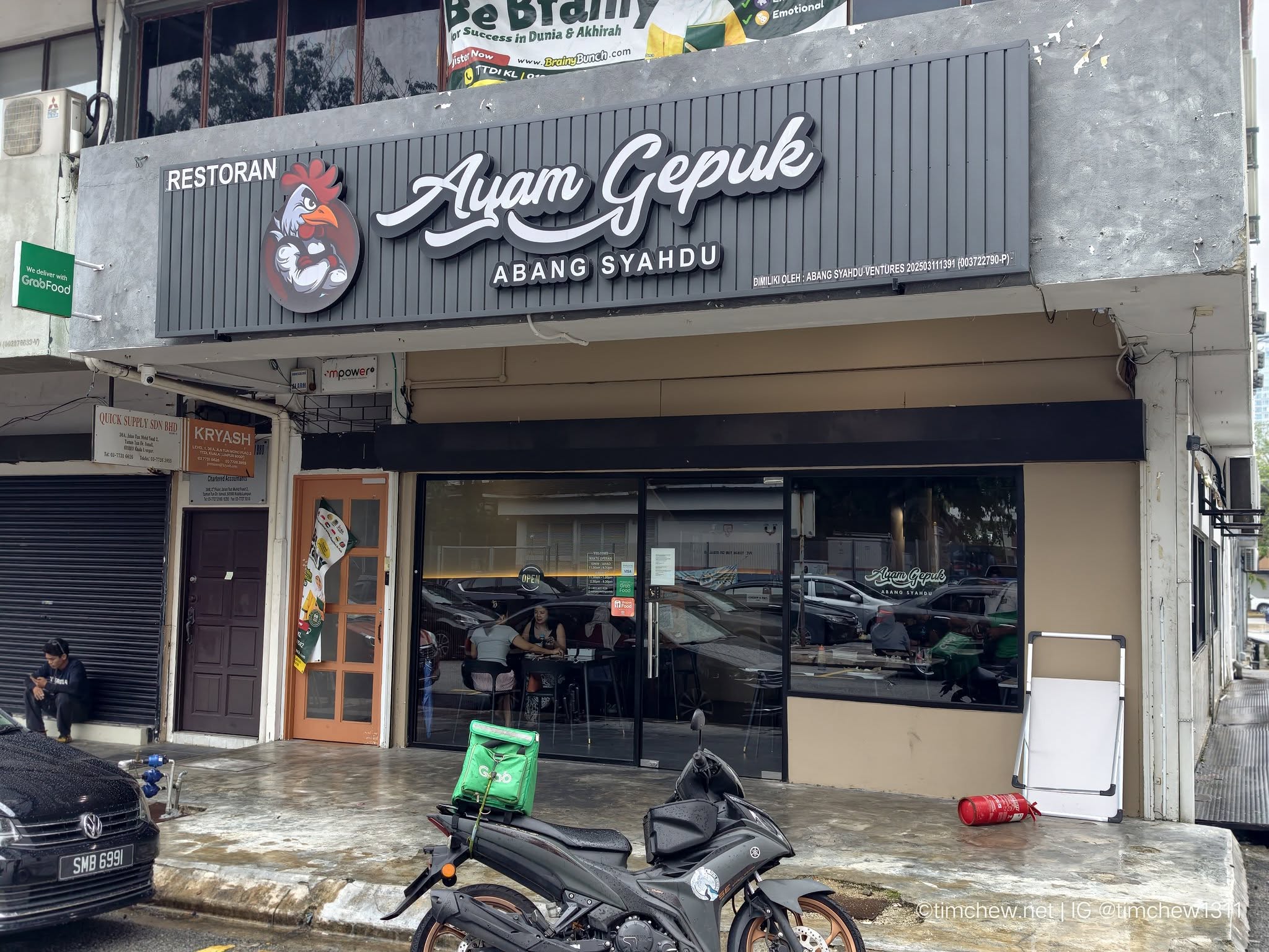 Exterior view of the restaurant 'Restoran Ayam Gepuk Abang Syahdu' featuring a prominent sign with a chicken logo. The space shows an open entrance with dining inside and a delivery sign for GrabFood visible.