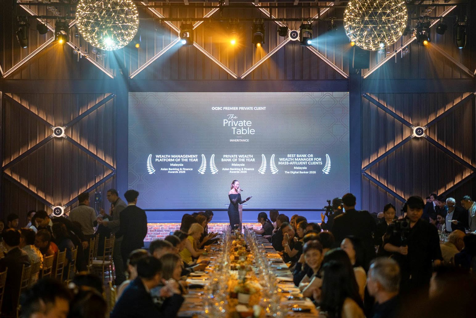 An elegant event setting with a long banquet table filled with guests, spotlighting a speaker in front of a large screen displaying awards for wealth management.