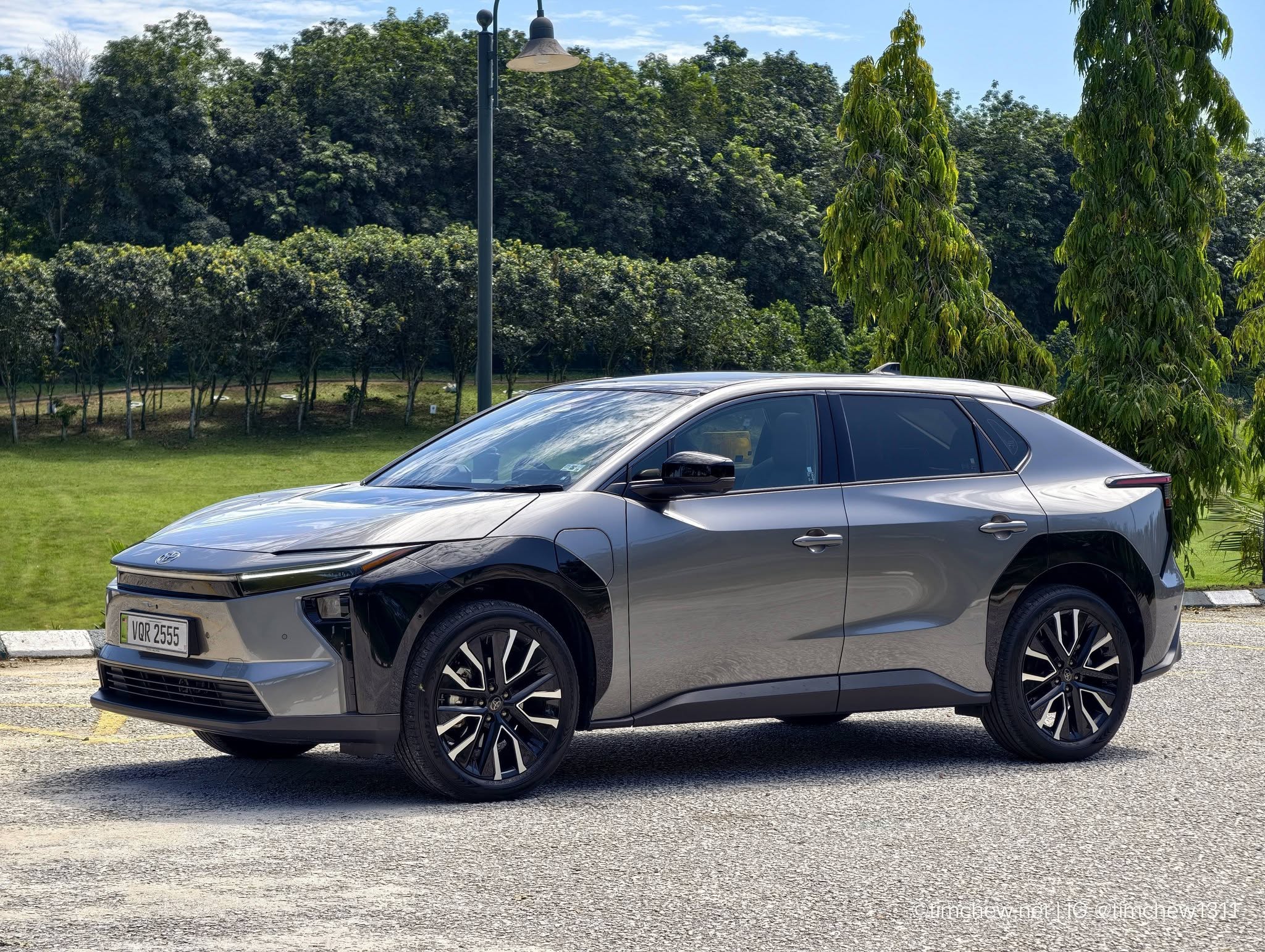A sleek gray electric car parked on a gravel surface, with greenery in the background and trees lining the scene.